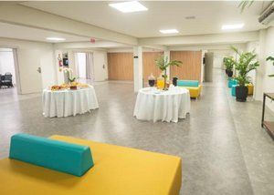 Salão de eventos luminoso com mesas redondas cobertas com toalhas brancas, bancos amarelos e almofadas verde-azuladas sob luzes de teto.