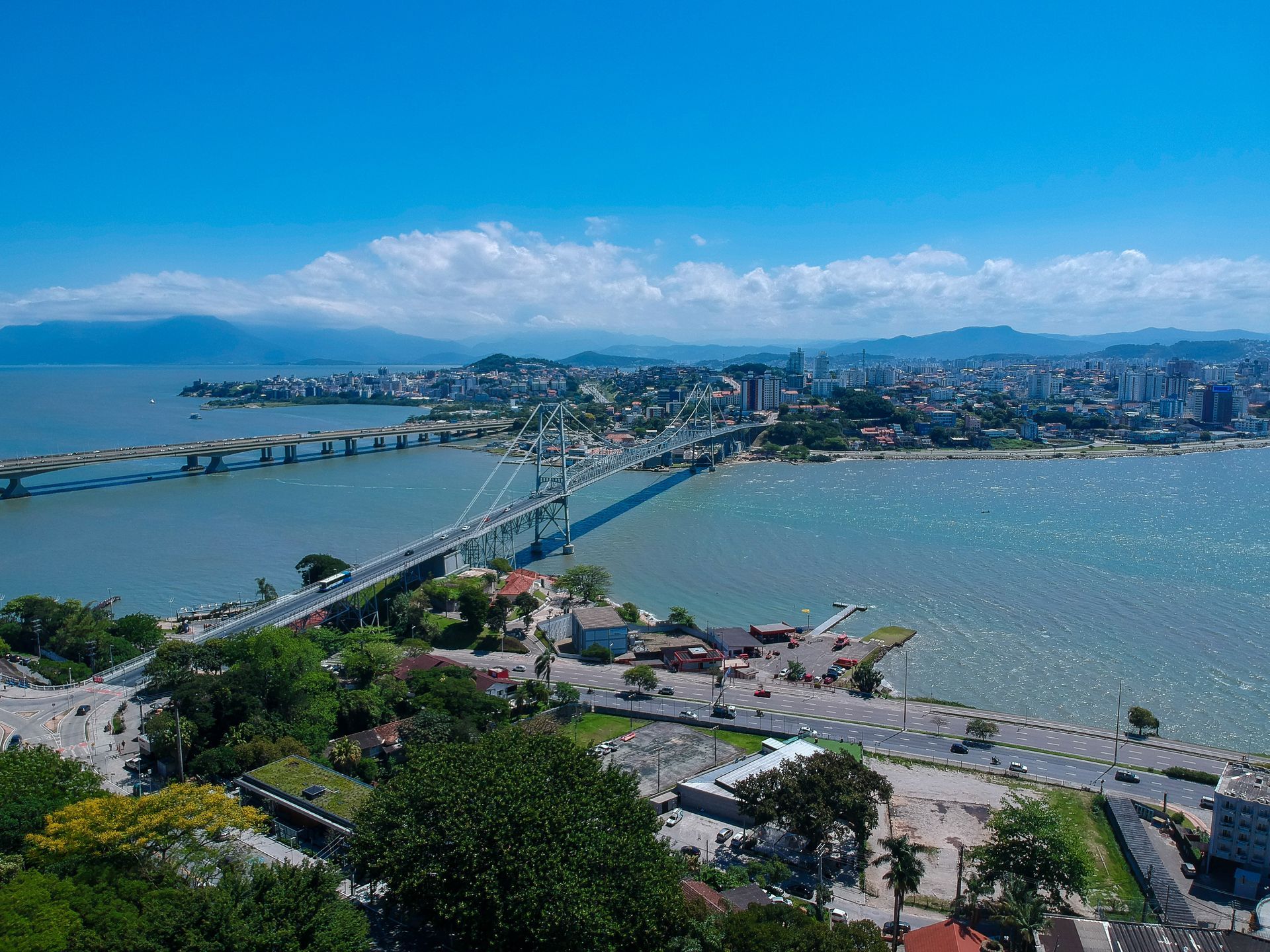 Ponte treliçada de aço sobre águas calmas ao lado de um prédio branco e margem gramada sob um céu azul claro.