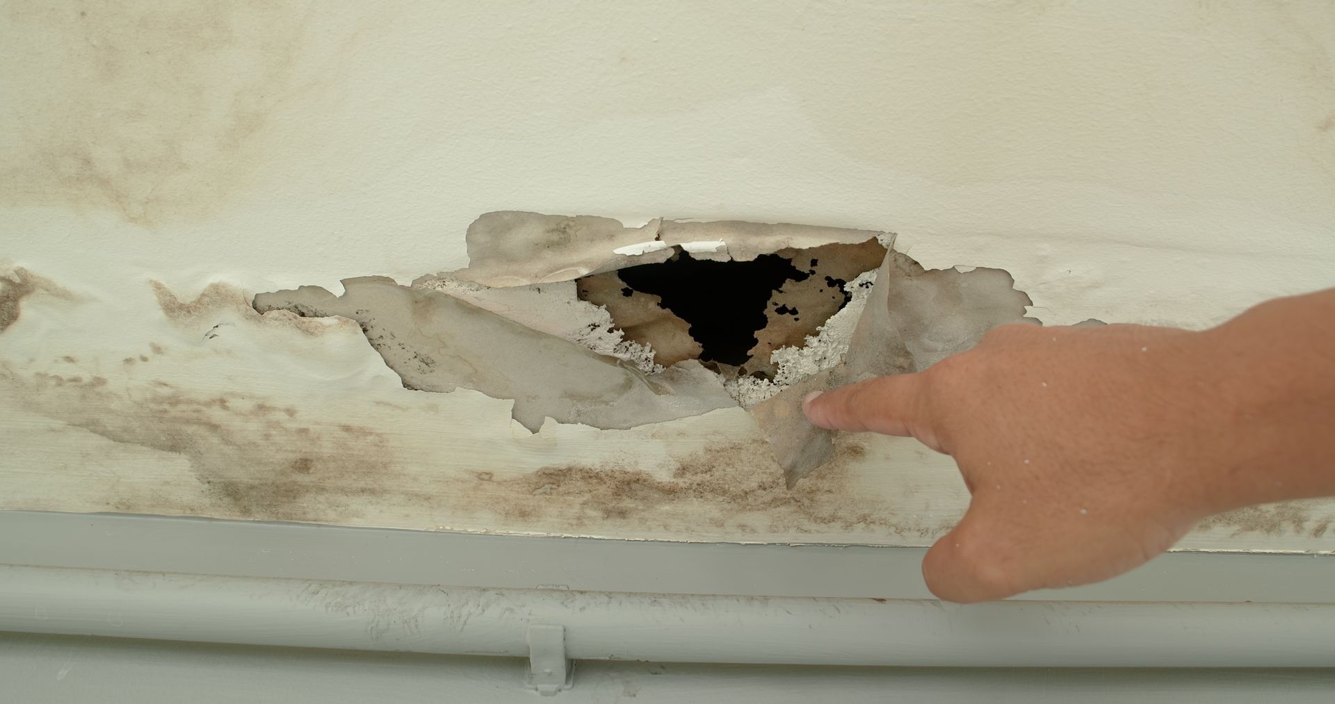 Hole in a ceiling with peeling paint, showing damage. A hand points at the hole.