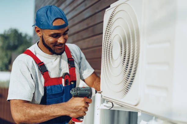 HVAC technician, smiling, wearing blue cap and overalls, working on an AC unit outside.
