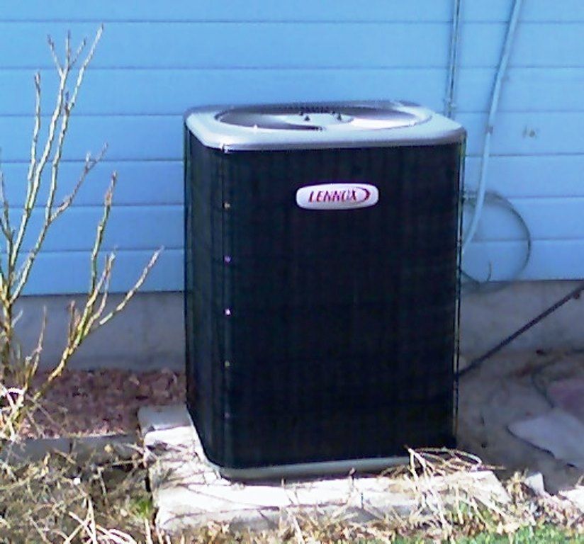 Lennox air conditioning unit outside a light blue house, near a bush and concrete slab.