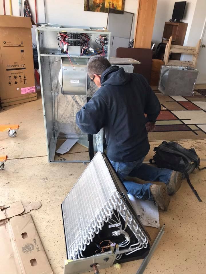 Man kneeling, working on HVAC unit in a garage. Metal components scattered; tools nearby.