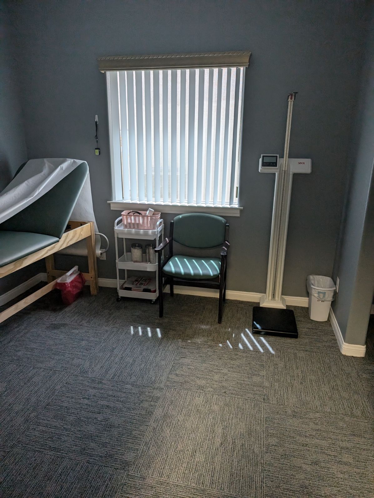 A doctor 's office with a bed , chair , scale and window.