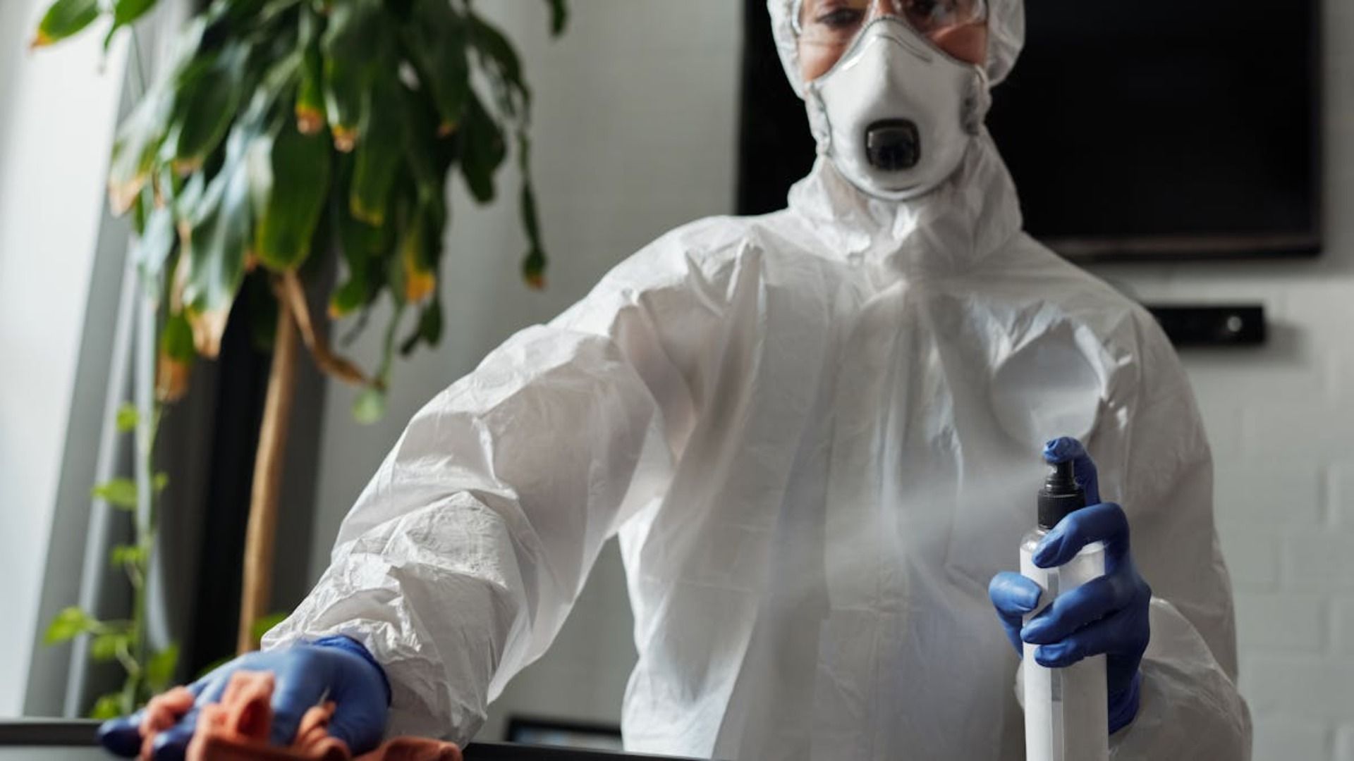 Persona con traje de materiales peligrosos desinfectando una superficie con una botella rociadora, en un lugar interior.