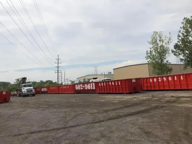 Red dumpsters lining a lot beside a beige building and a parked truck under cloudy skies.