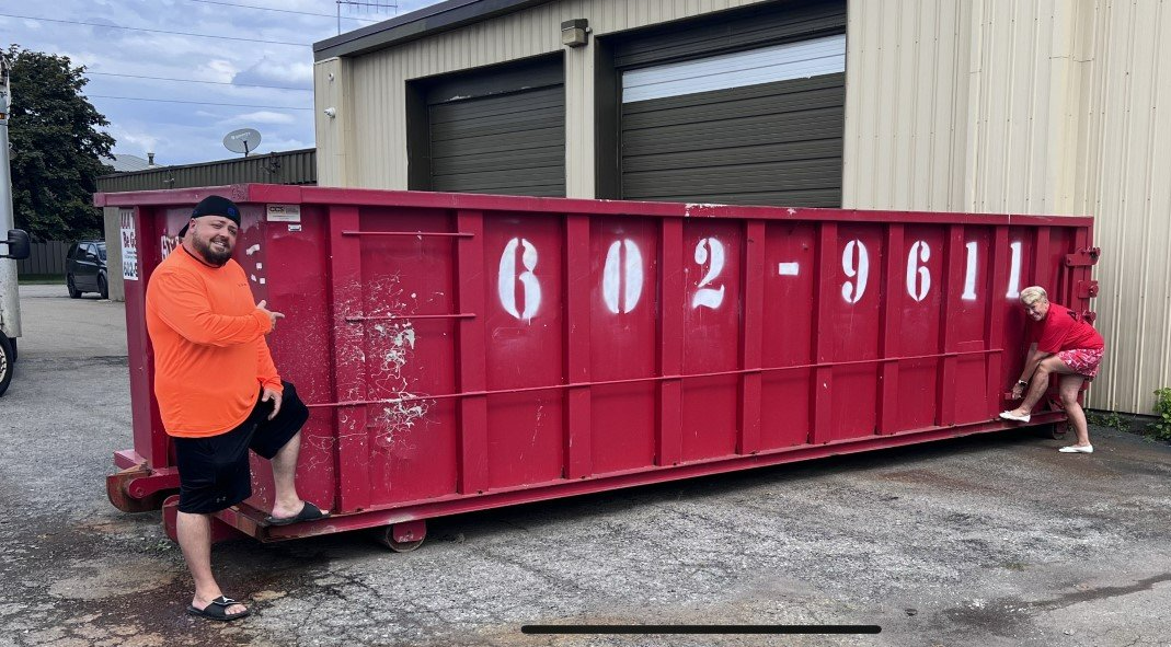 Red dumpster in a gravel lot beside a garage, with two people standing at the ends and white numbers on the side