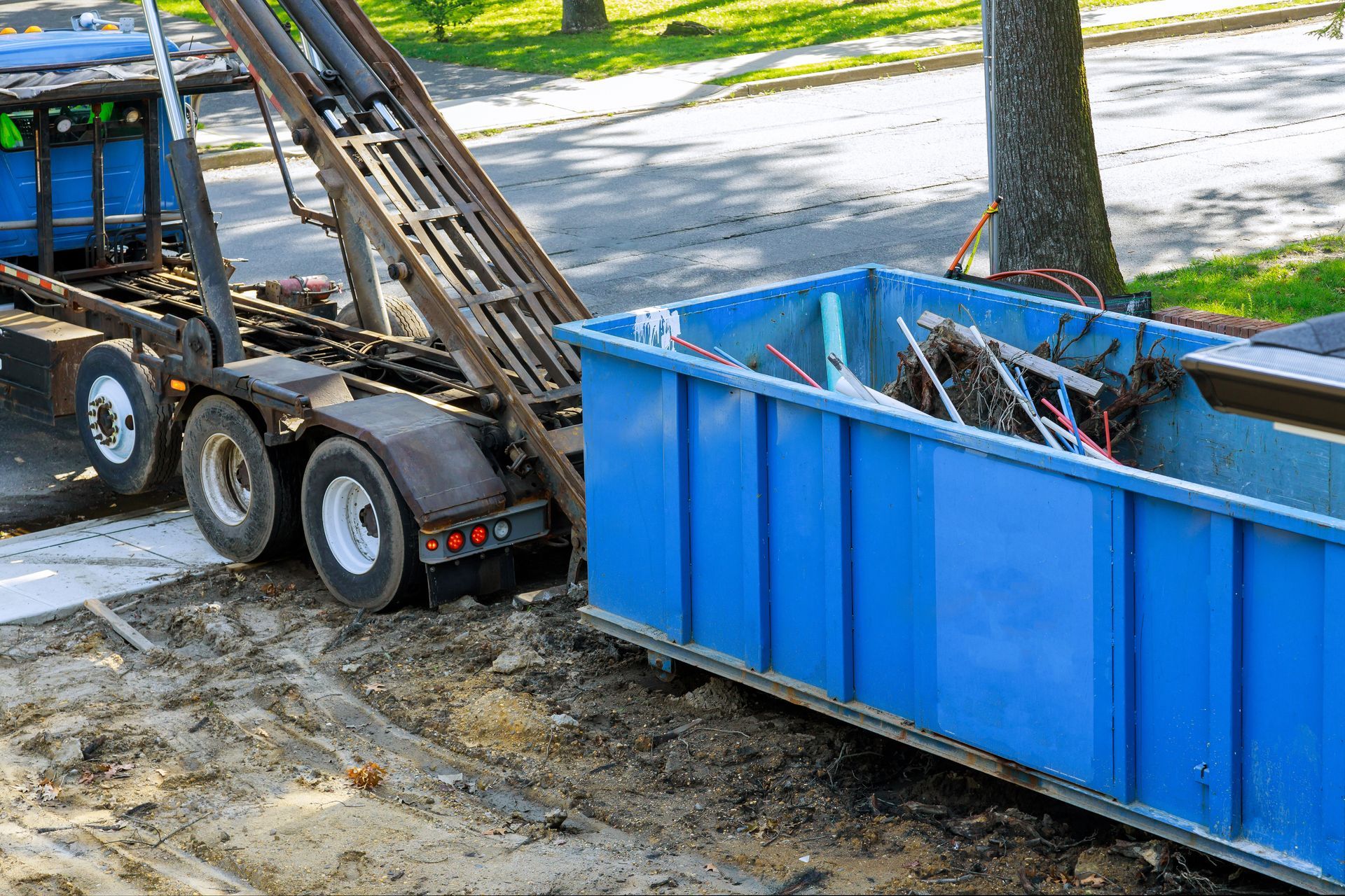 Blue roll-off dumpster positioned beside a truck, loaded with debris and scrap materials.