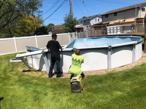 Two workers repairing an above-ground pool cover in a backyard with a fence and houses nearby