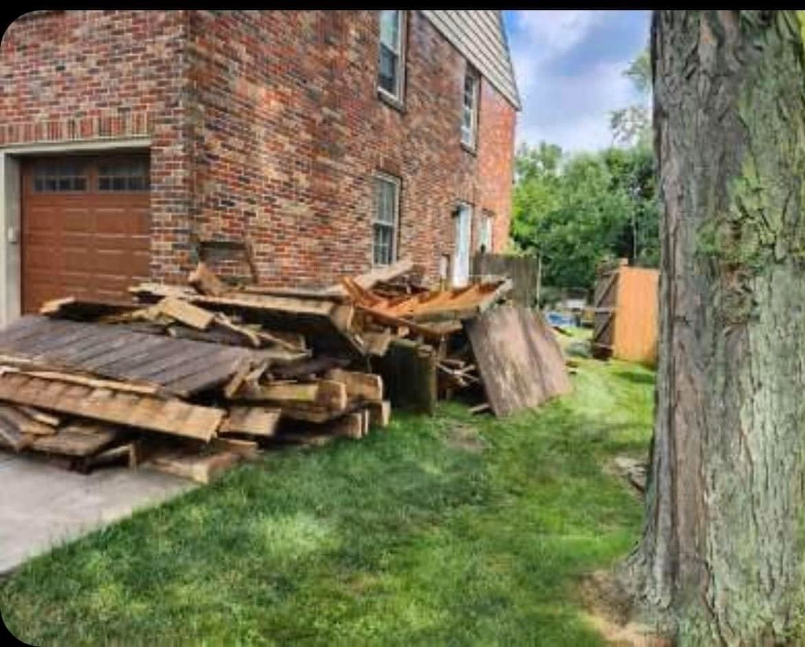 Piled wooden debris beside a brick house next to a grassy yard and tree trunk