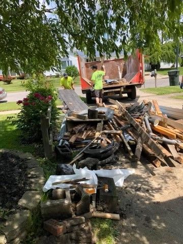 Workers loading debris and lumber into a truck beside a pile of construction waste on a street under trees