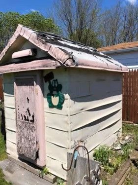 Small weathered shed with a slanted roof and green graffiti, beside a fence and trees.