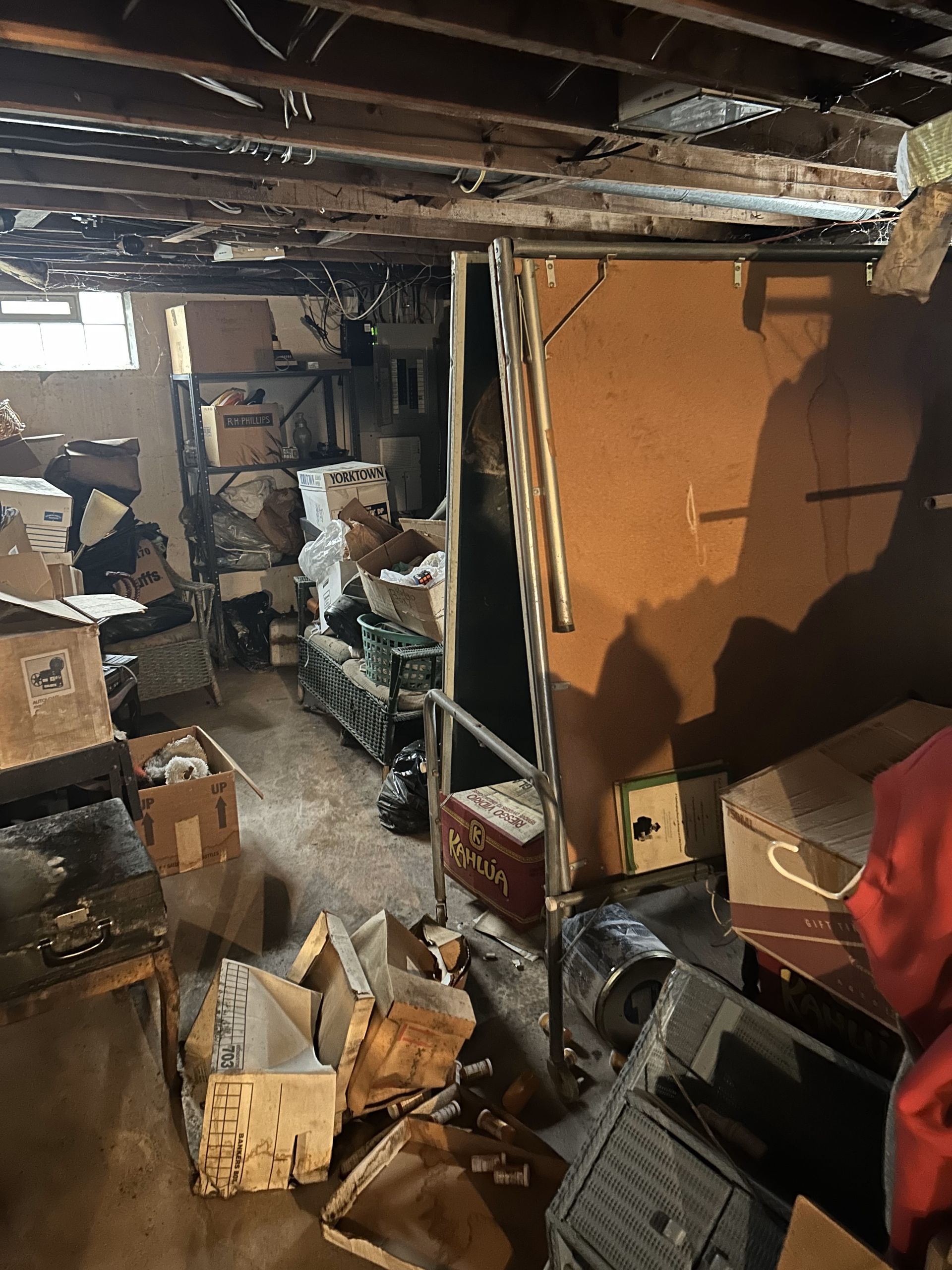 Cluttered basement with boxes, debris, and damaged walls under exposed ceiling beams