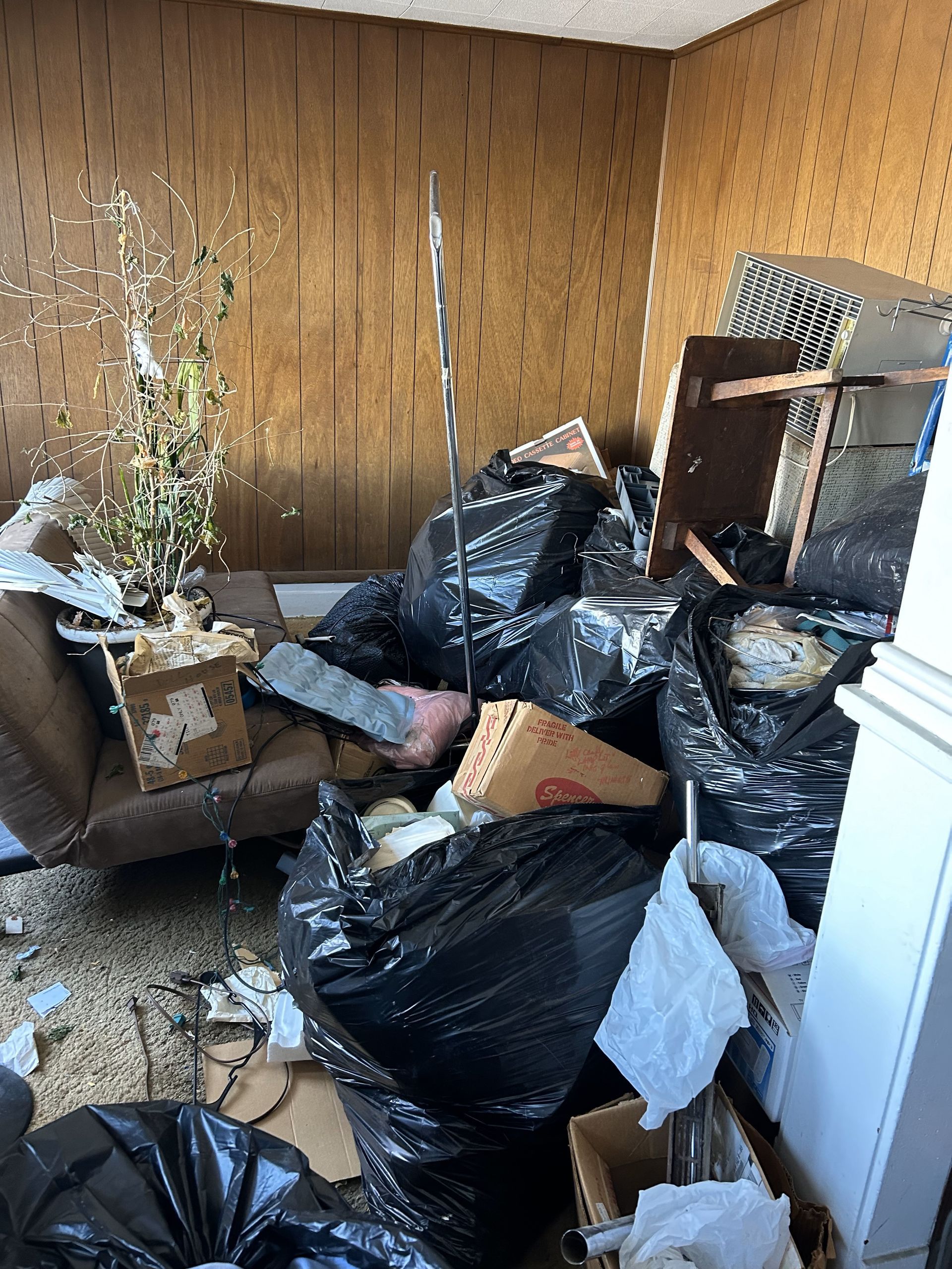 Cluttered room with overflowing black trash bags, boxes, and debris against wood-paneled walls.
