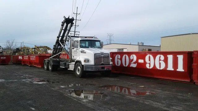 Tow truck in a lot beside a red dumpster with large white numbers, under a cloudy sky