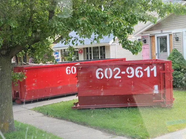 Two red dumpsters on a suburban driveway in front of a house, one marked “602-9611”