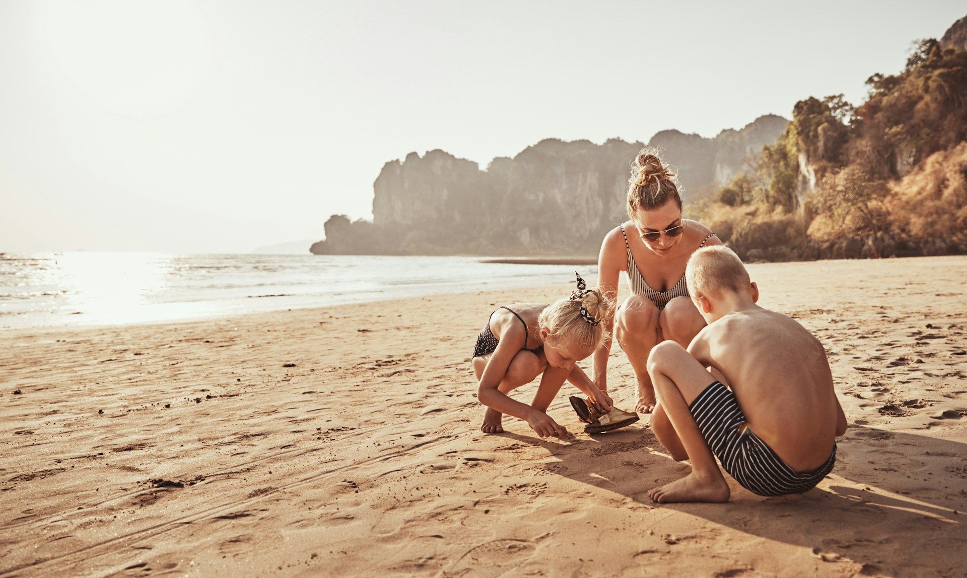 A woman and two children play on a sandy beach, mountains in the background.