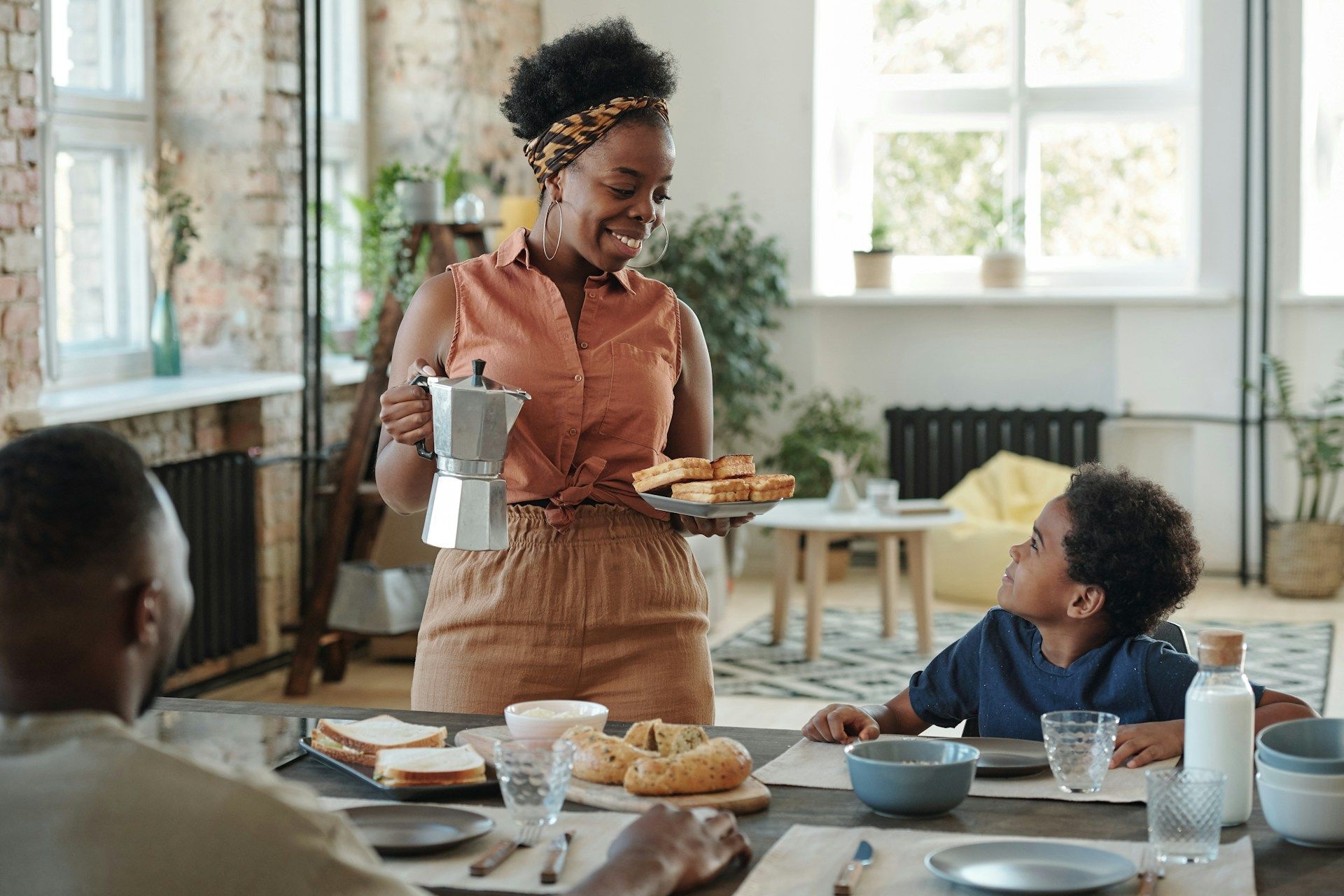 Woman serving breakfast to family: holding coffee pot, pastries; dining table setting.