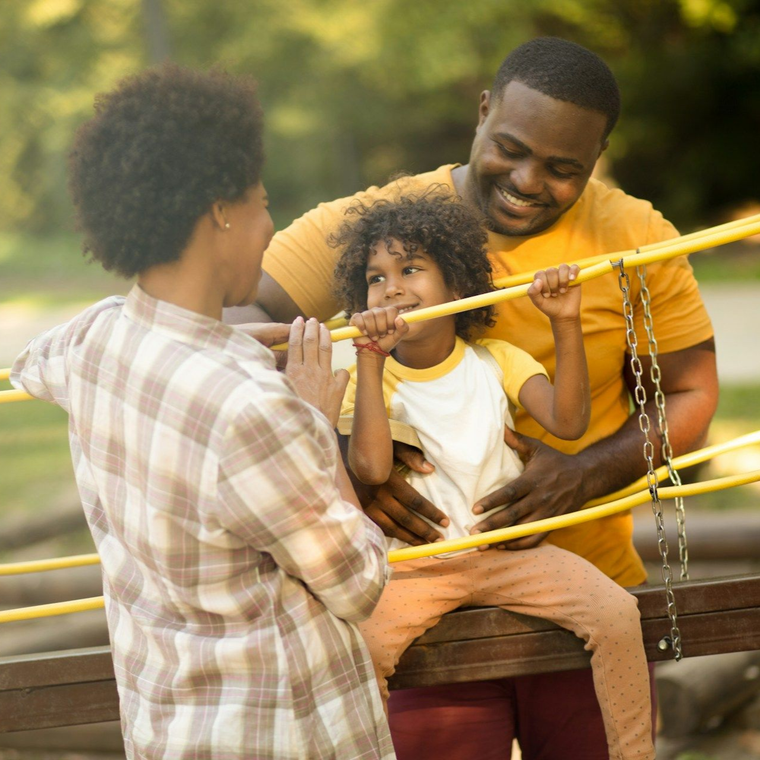 Family at a park: a Black mother and father smile at their child playing on a yellow structure.