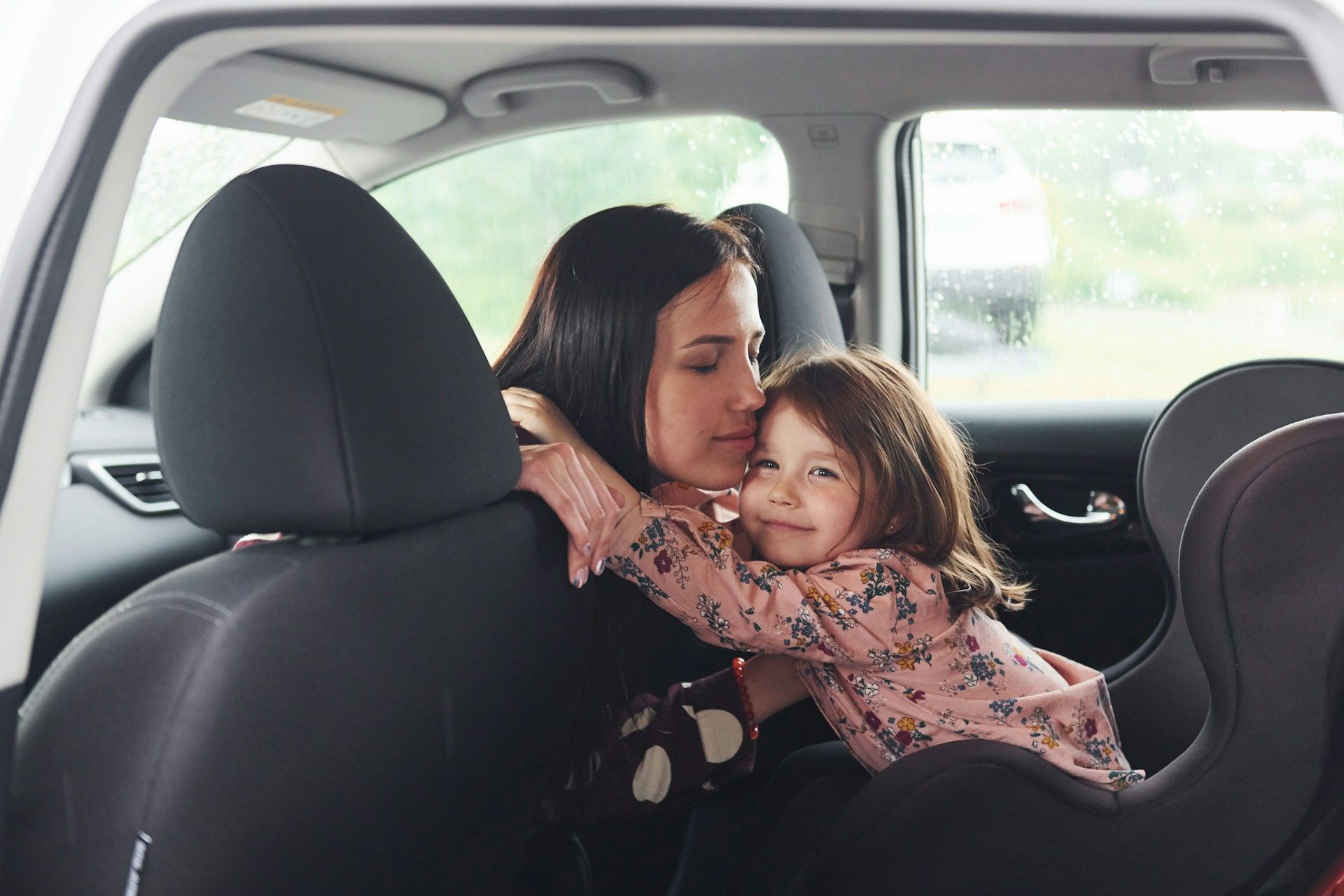 Woman hugs a young girl sitting in a car seat inside a car. The woman looks down smiling.