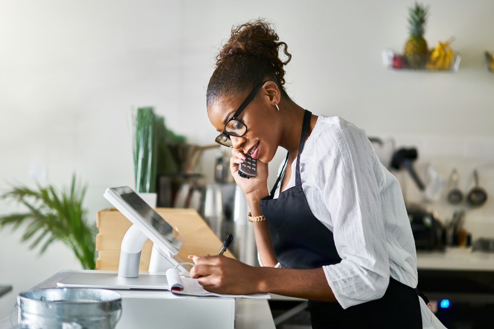 Woman in apron taking phone order, writing on notepad, looking at tablet in a cafe.