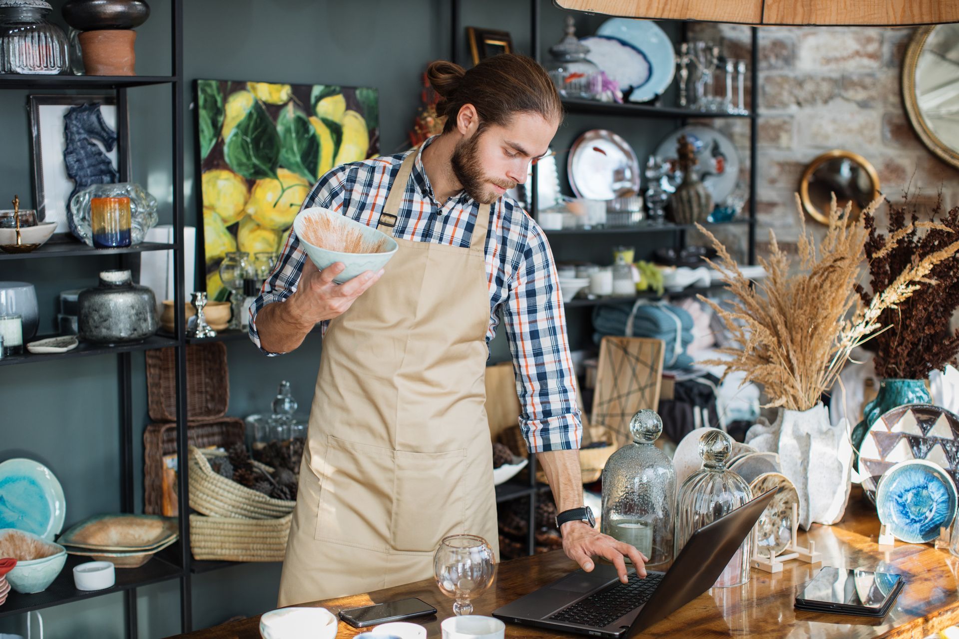 Man in apron working on laptop in a shop, holding a bowl, surrounded by shelves of goods.