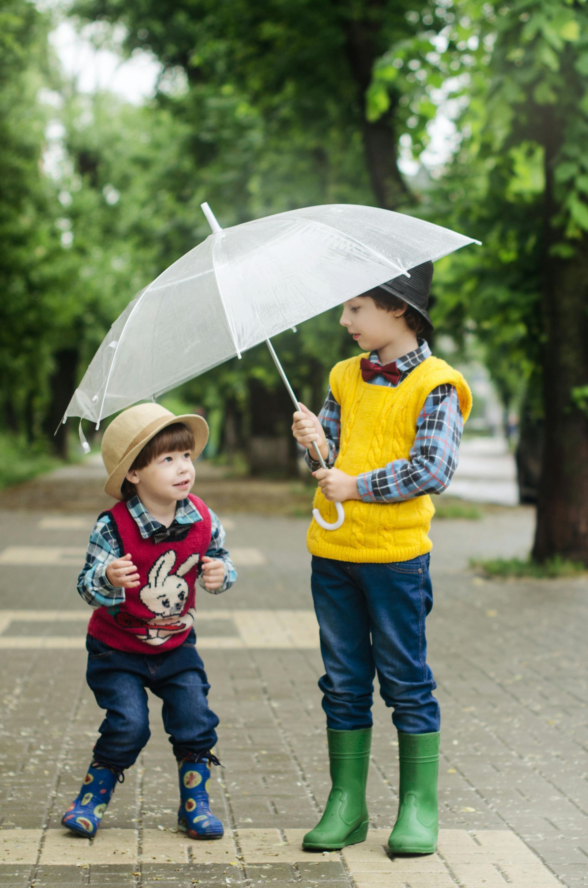 Two young boys in rain gear under an umbrella on a brick walkway.