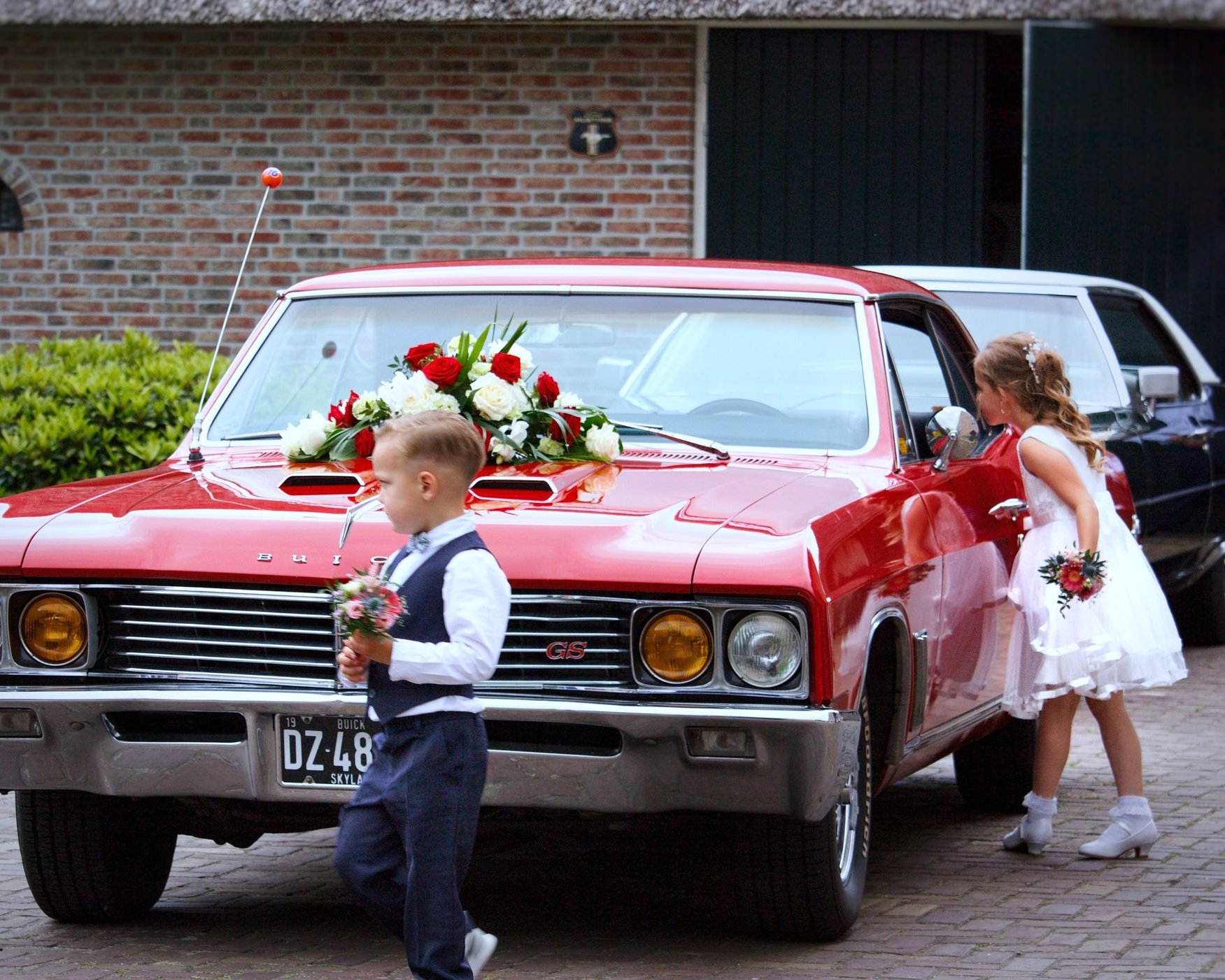 Een rode klassieke auto versierd met bloemen, twee kinderen bij de auto, waarvan er één bloemen vasthoudt.