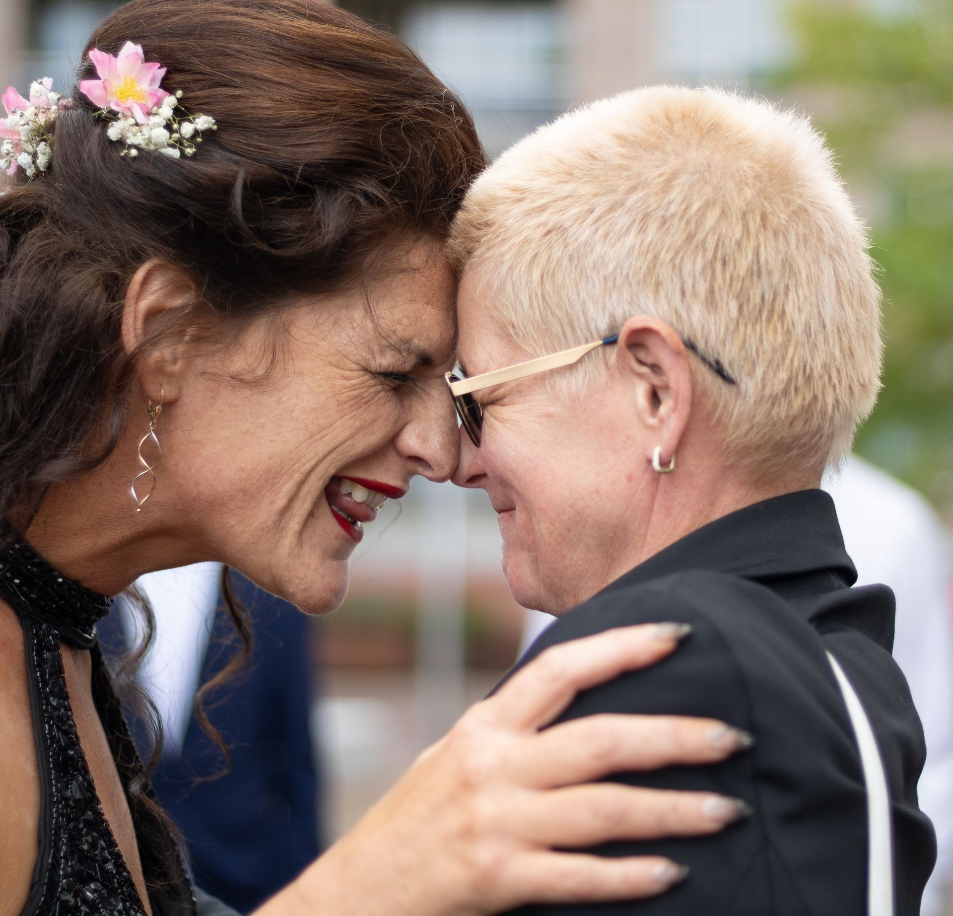 Twee mensen raken elkaars voorhoofd aan en glimlachen. De ene heeft donker haar en bloemen, de andere kort blond haar en een bril.