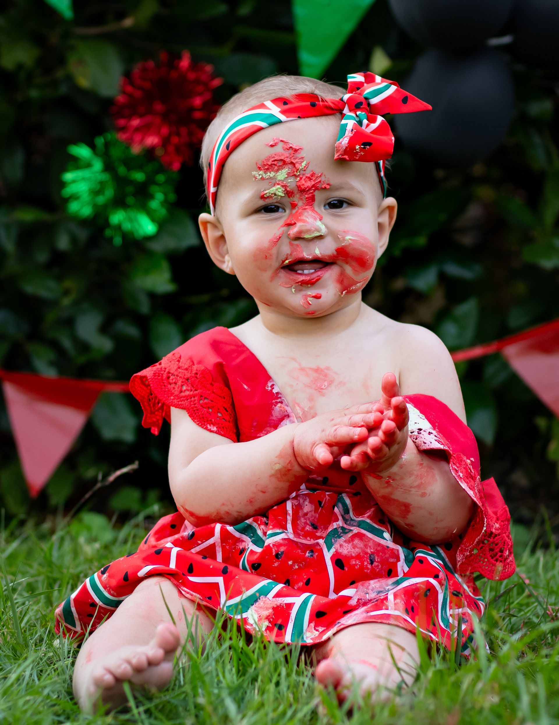 Een baby, bedekt met rode taart, met een jurkje en hoofdband met watermeloenprint aan, zit lachend in het gras.