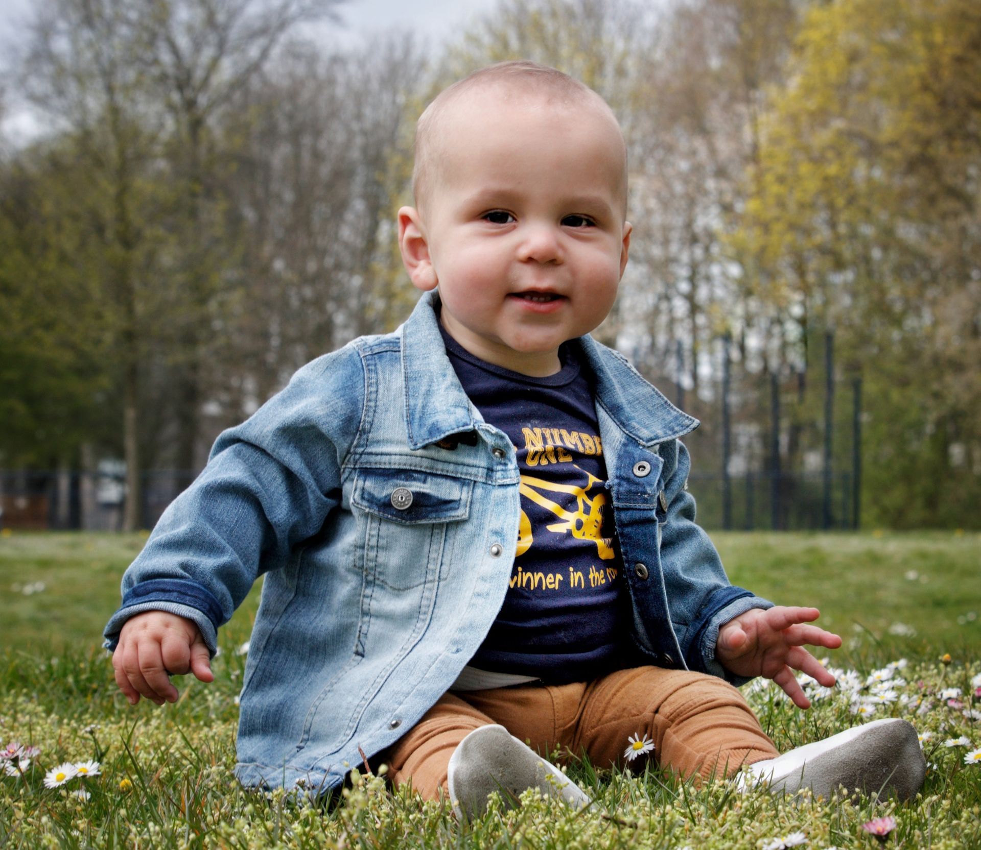Een baby, gekleed in een blauw spijkerjasje en een bruine broek, zit lachend in het gras.