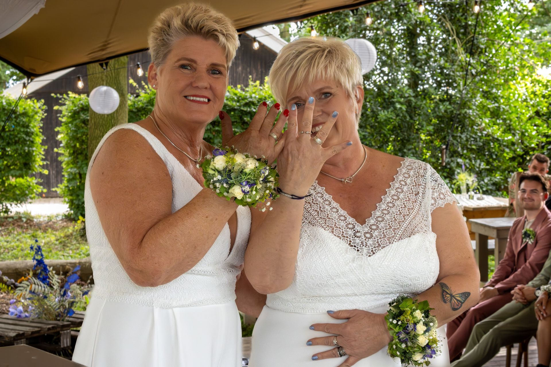Twee vrouwen in witte jurken vieren een bruiloft in de buitenlucht en tonen hun ringen.