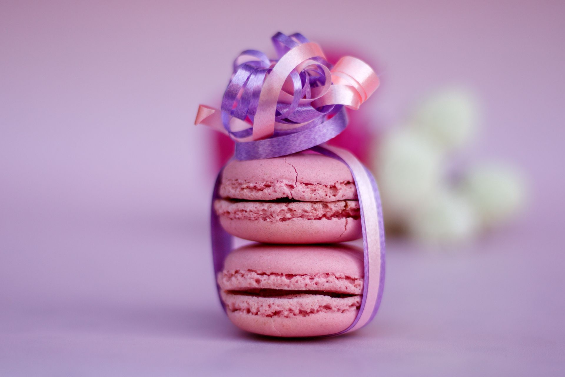 Twee roze macarons op elkaar gestapeld en vastgebonden met lavendelkleurig en roze lint, op een roze achtergrond.