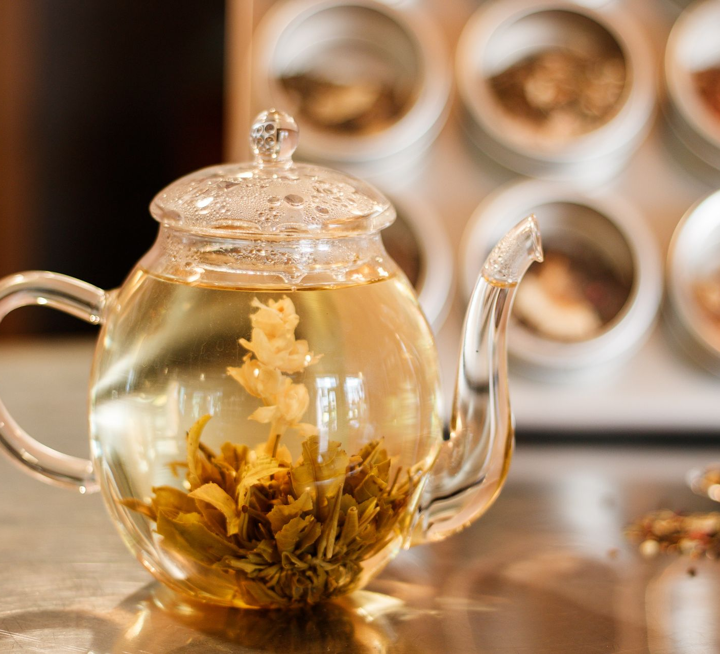 Glazen theepot met bloeiende theebloemen, op een reflecterend oppervlak, met theepotten op de achtergrond.