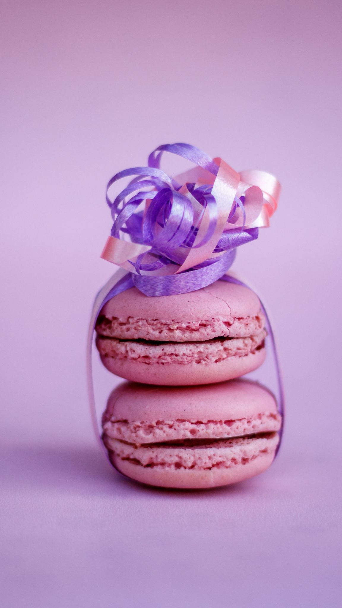 Twee roze macarons, vastgebonden met een paars en perzikkleurig lint, op een lila achtergrond.