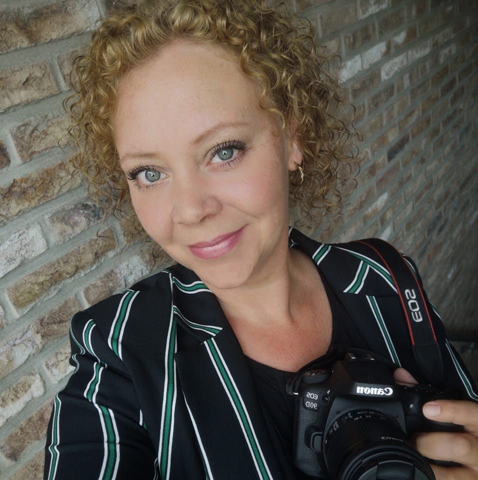 Vrouw met krullend blond haar, een gestreepte blazer, een Canon-camera in haar hand, lachend bij een bakstenen muur.