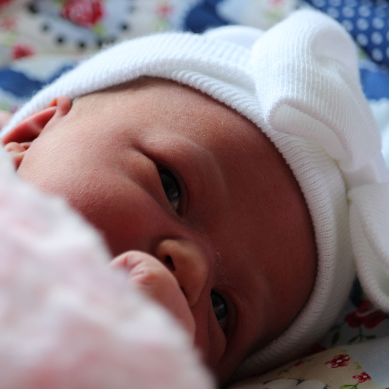 Pasgeboren baby met wit mutsje op, kijkt omhoog met zachte huid en een lichte glimlach.
