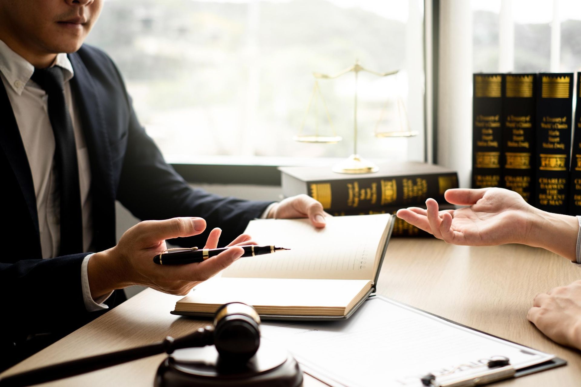 Lawyer in suit discussing notes with a client; gavel, books, and scale of justice on desk.