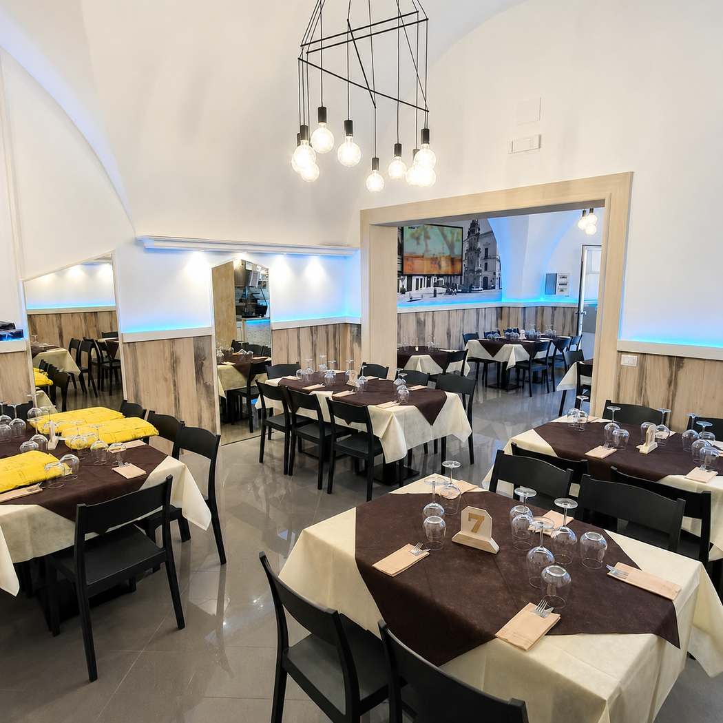 Sala da pranzo vuota di un ristorante con tavoli neri, sedie beige, pareti bianche e lampade a sospensione.