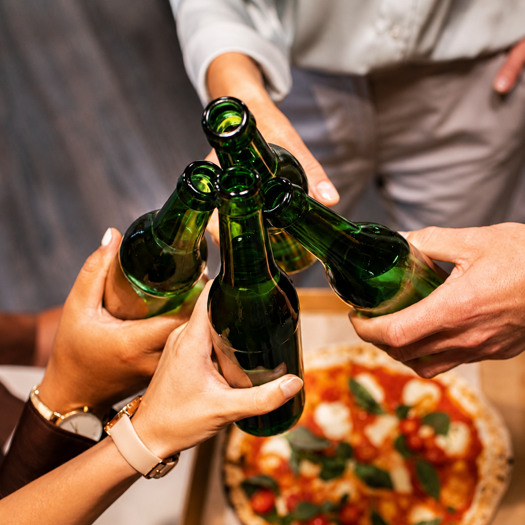 Mani che tintinnano bottiglie di birra verde sopra una pizza a un tavolo.