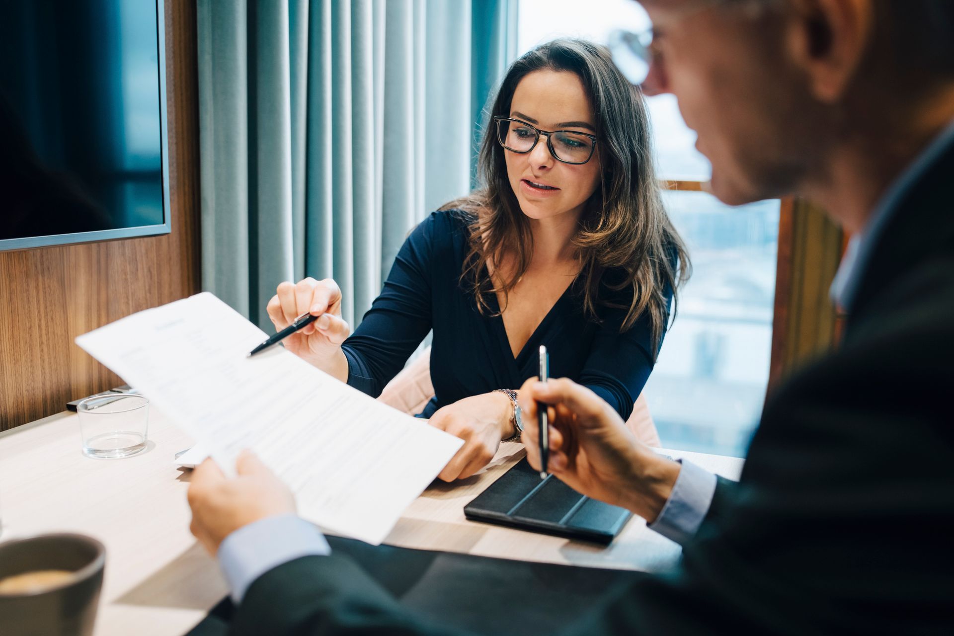 Woman in glasses points at document, discussing with a man at a desk.