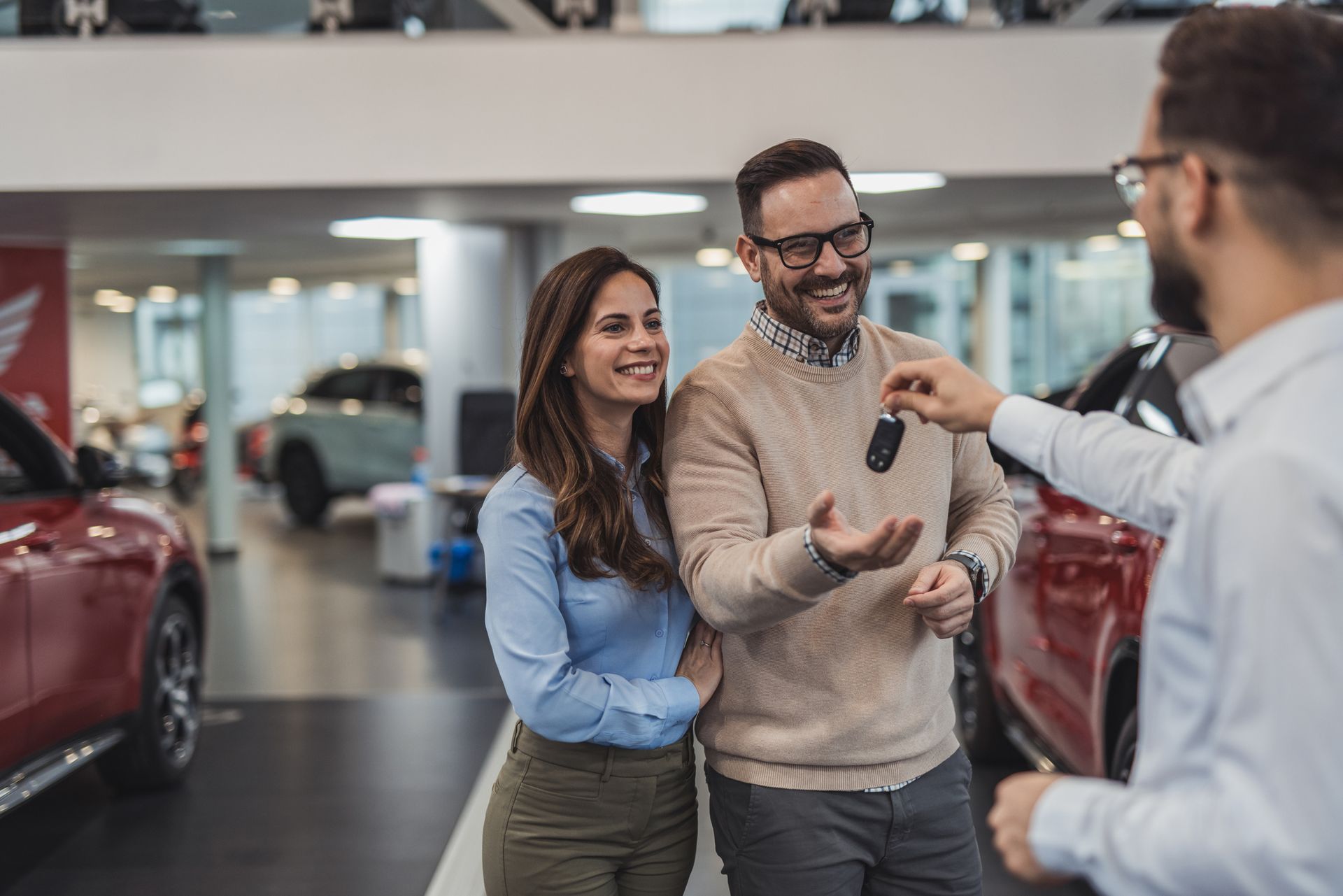 Couple receives car keys from a car dealer at a dealership; they are smiling.