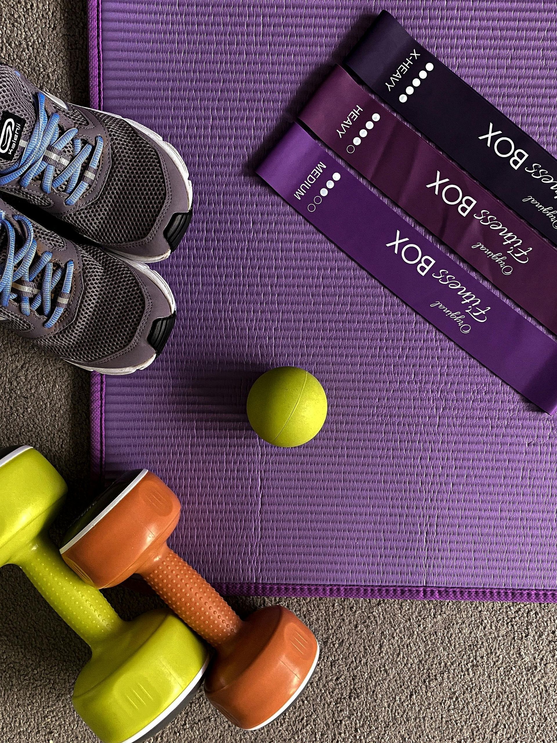 Workout gear on purple mat: running shoes, dumbbells, resistance bands, and a tennis ball.