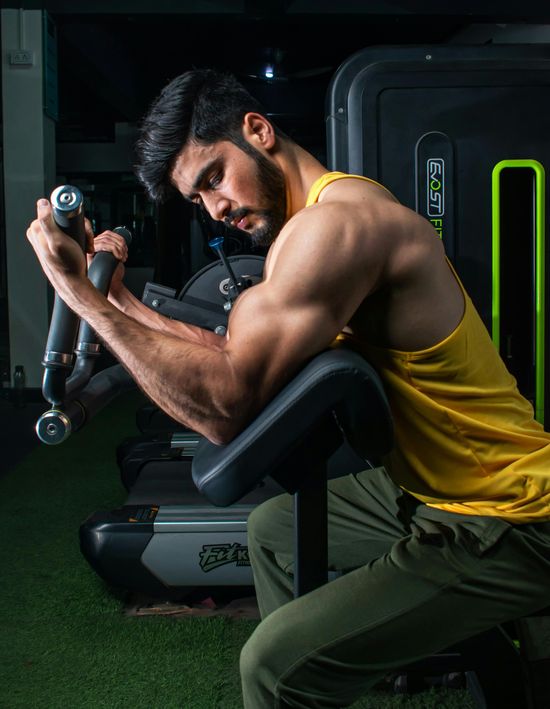 Man using a dumbbell preacher curl on a gym bench, wearing a yellow tank top and green pants.
