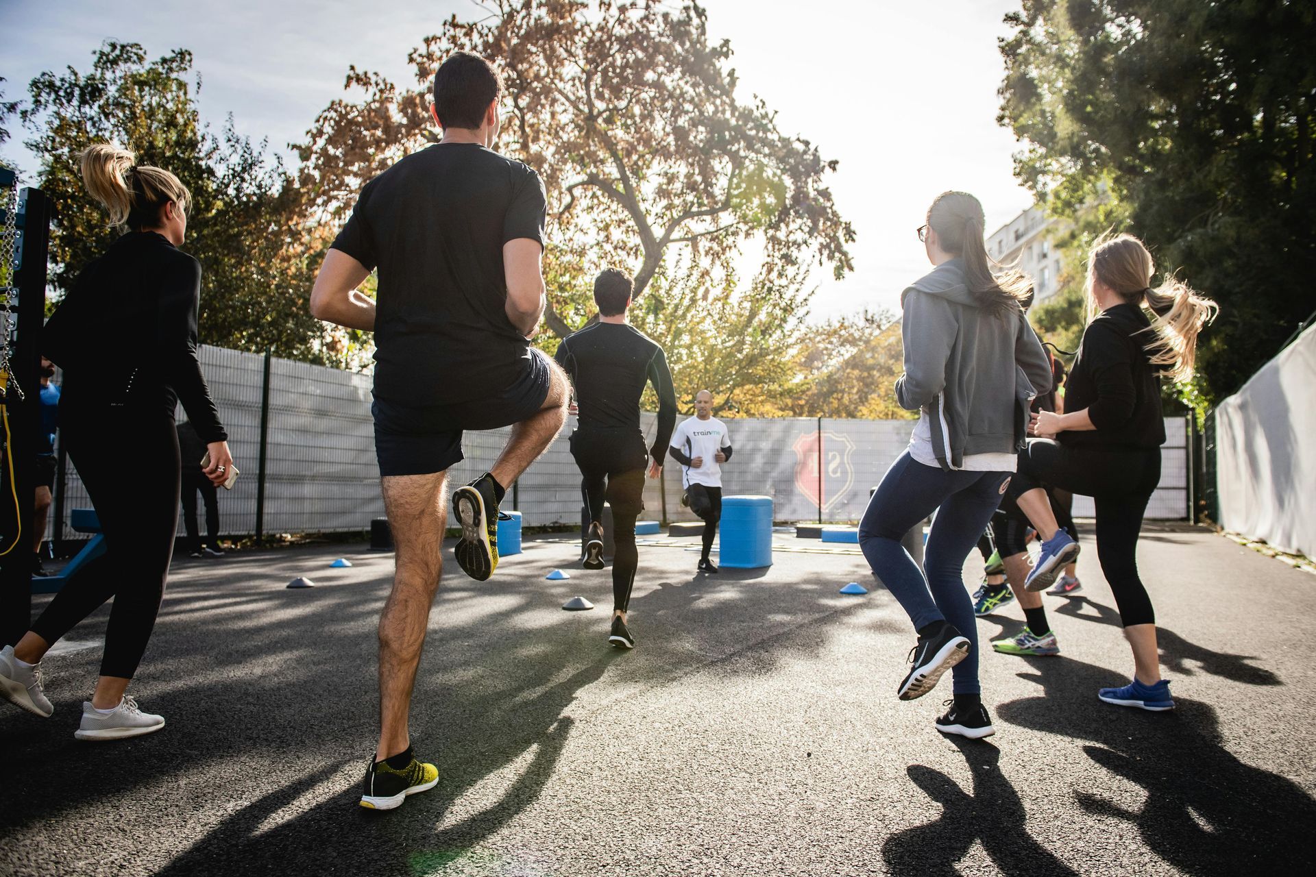 People exercising outdoors on a sunny pavement, stretching and jogging near a white wall and trees.