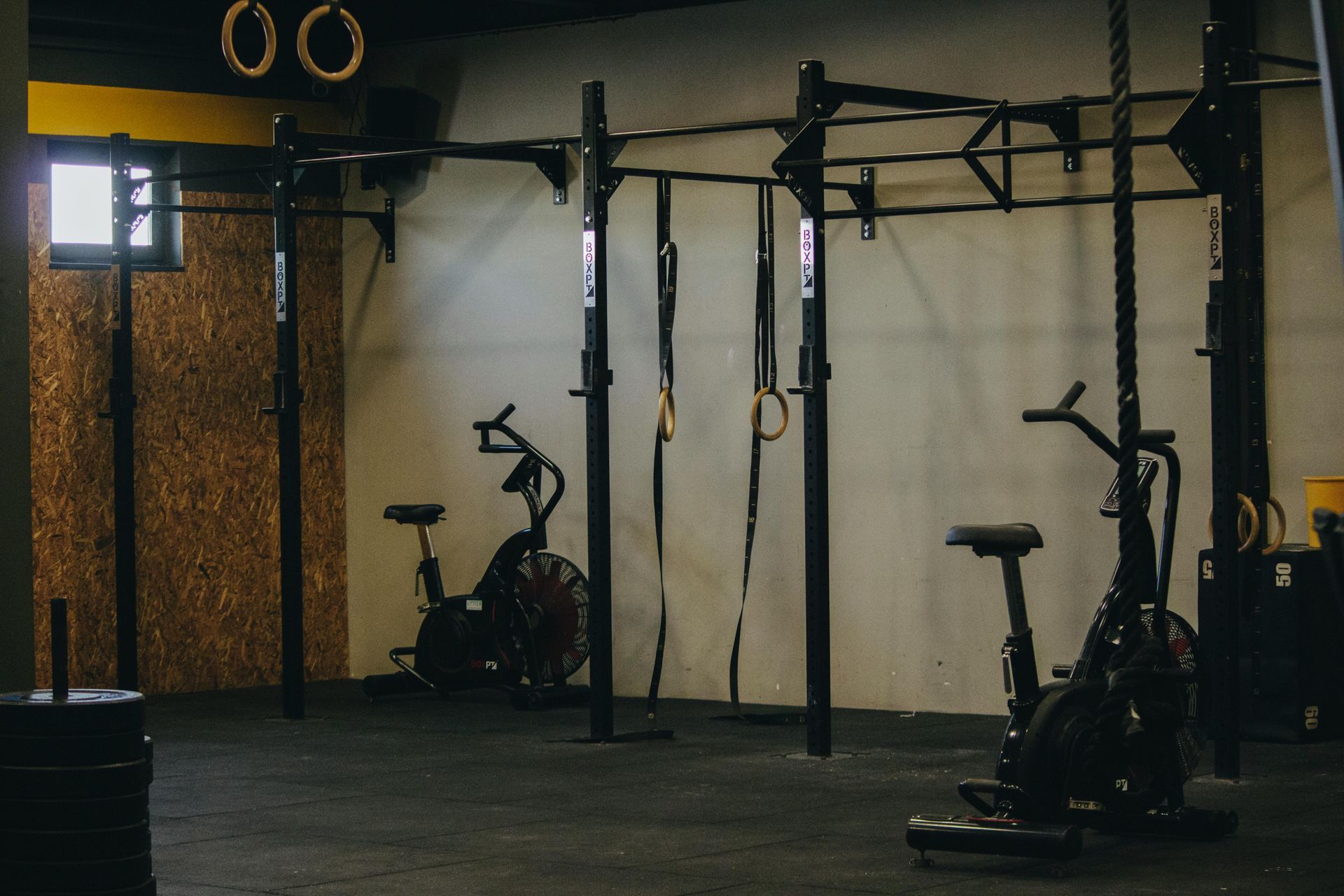 Dim gym with stationary bikes, pull-up rig, and gymnastic rings on a dark floor