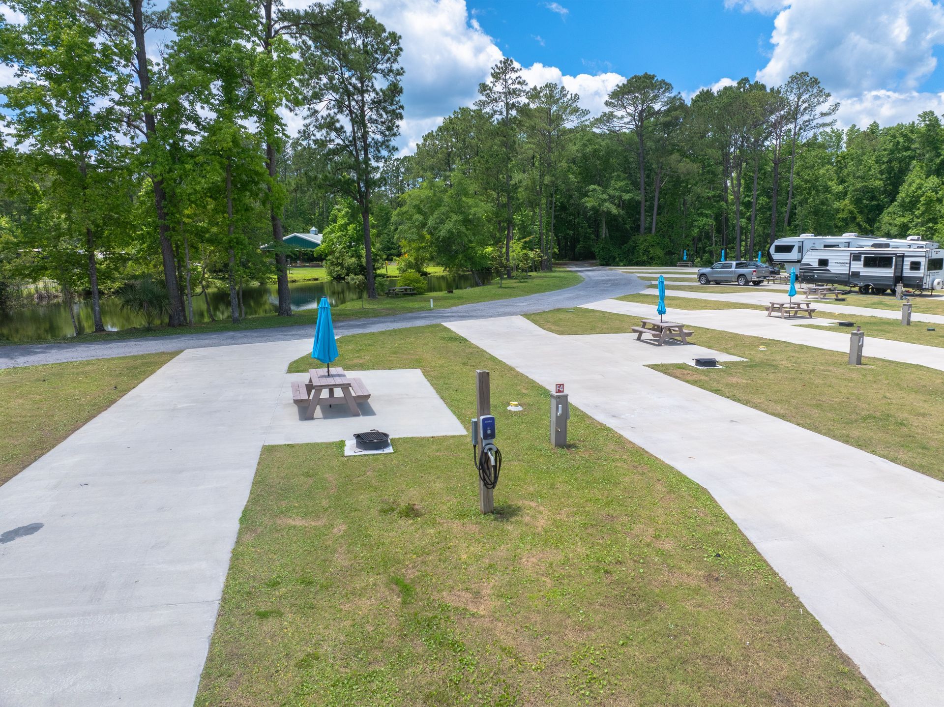 RV park with concrete pads, picnic tables, and RVs parked, surrounded by trees.
