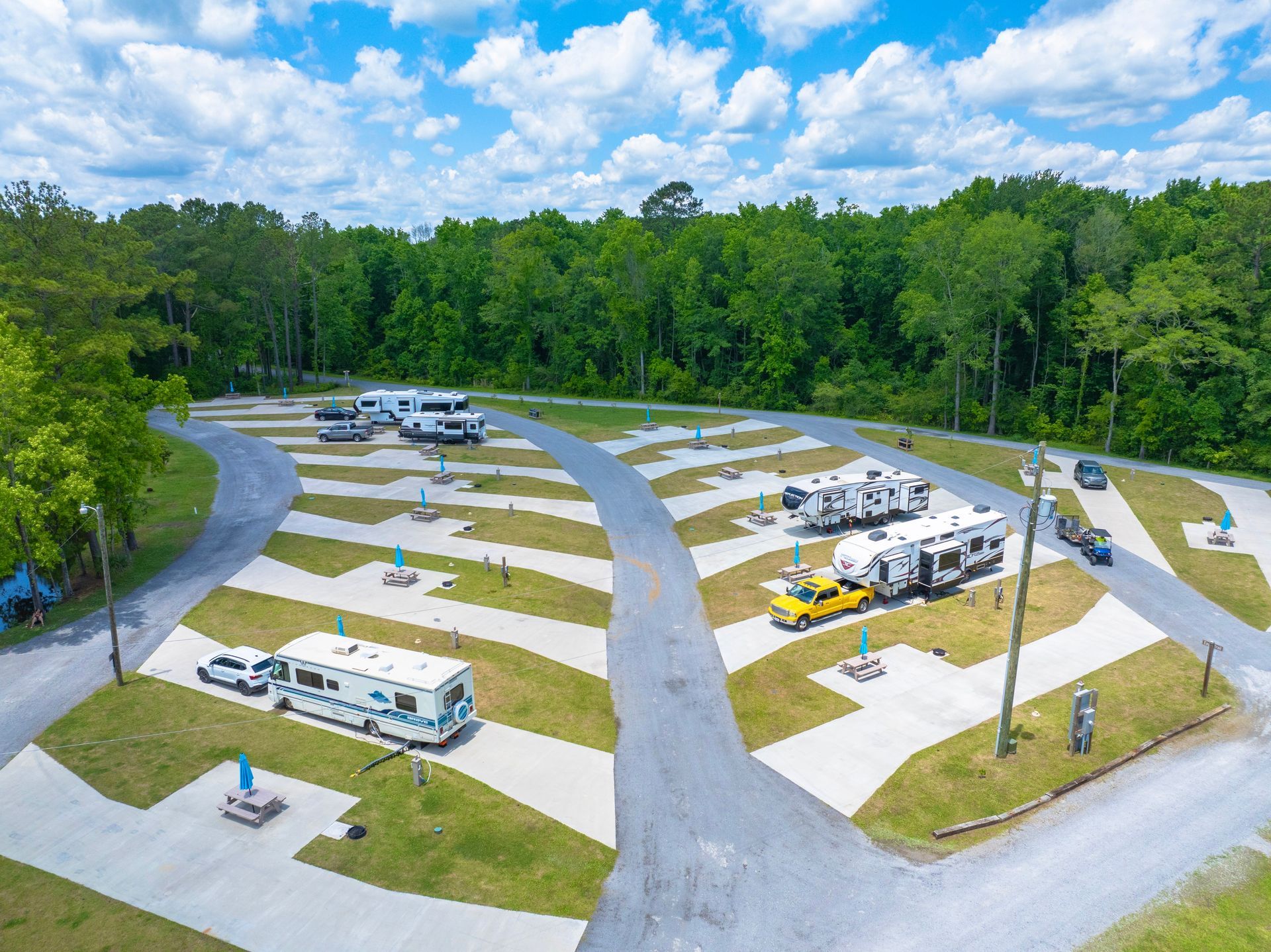 Aerial view of RV campground with parked RVs on paved sites, green grass, and trees.