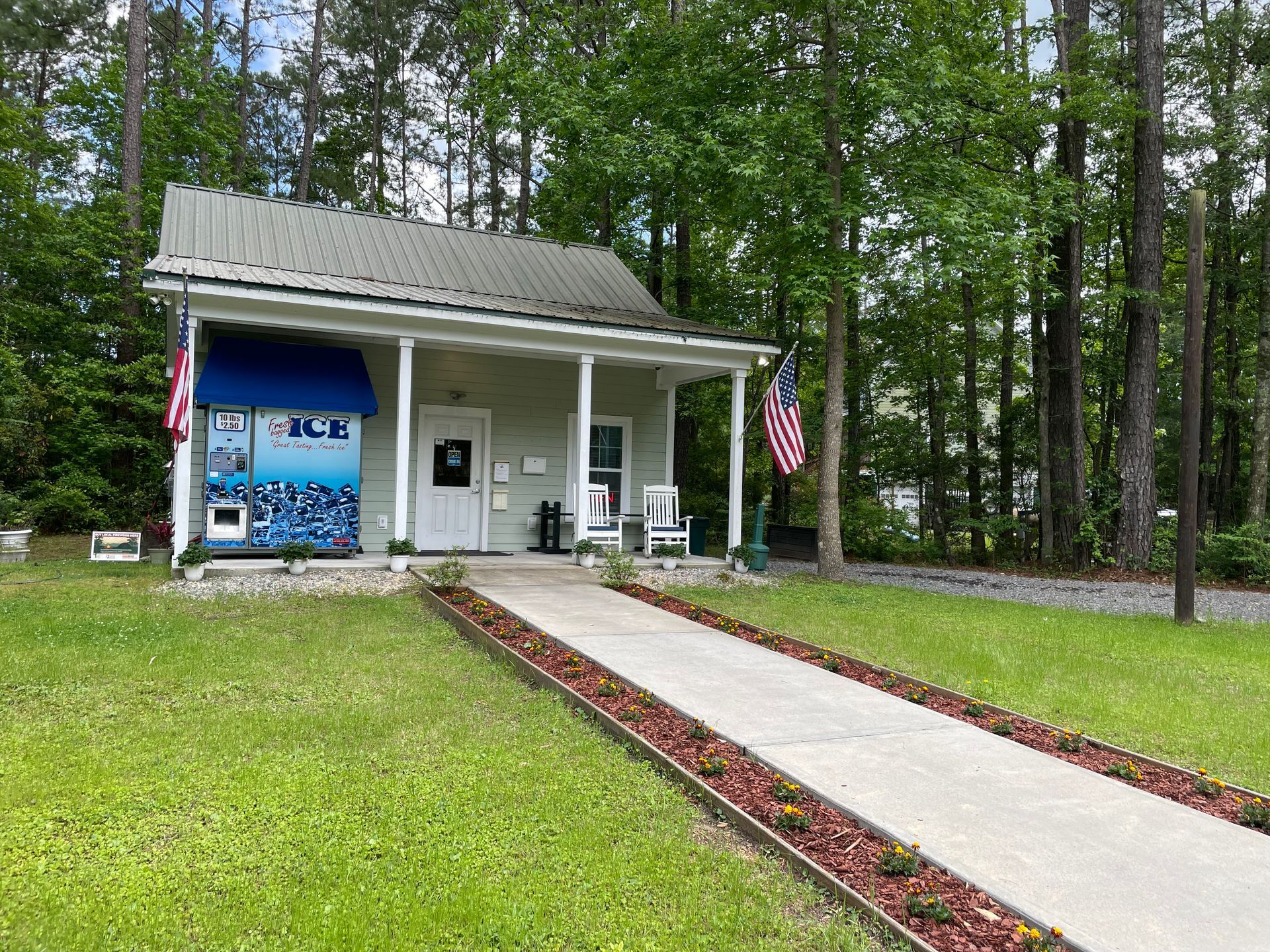 Small gray building with porch, ice machine, walkway, and American flags in a grassy area.
