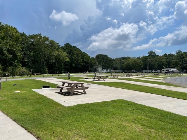A campground with concrete pads, picnic tables, and green grass. Forest in the background, blue sky with clouds.