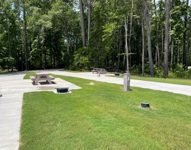 Campground with picnic tables, grassy areas, and tall trees. Paved road and utilities visible.